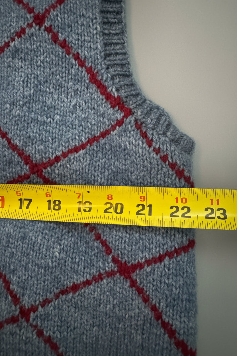 Close-up of a blue knit sweater with a red argyle pattern, shown next to a beige garment and a yellow measuring tape across the front.