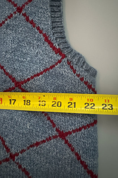 Close-up of a blue knit sweater with a red argyle pattern, shown next to a beige garment and a yellow measuring tape across the front.