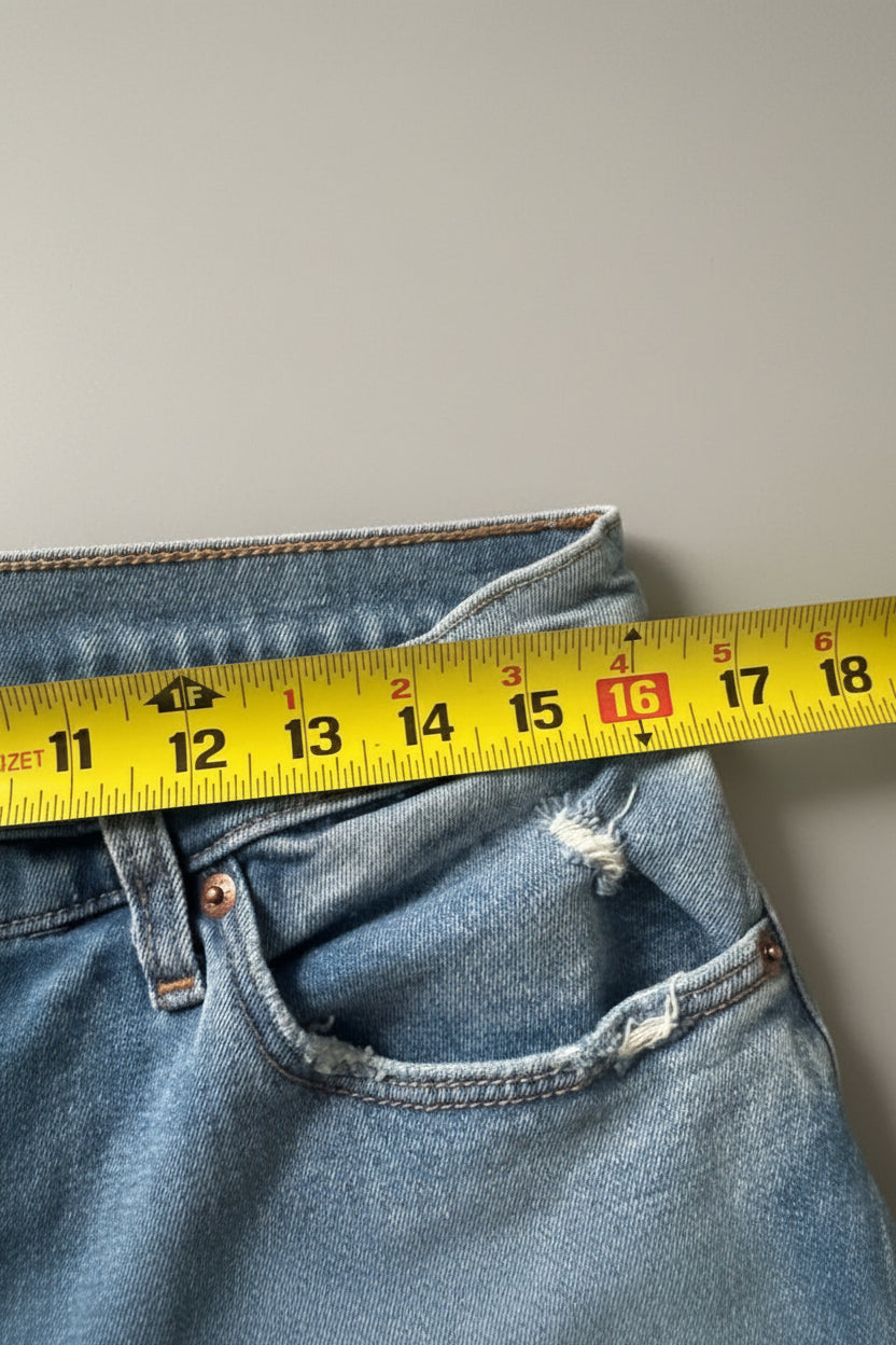 Close-up of blue denim jeans laid flat on a textured beige fabric surface, with a yellow measuring tape ruler placed horizontally across the front pocket area to show dimensions.