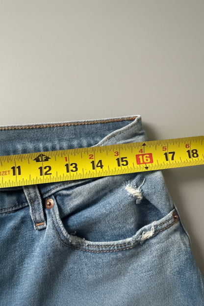 Close-up of blue denim jeans laid flat on a textured beige fabric surface, with a yellow measuring tape ruler placed horizontally across the front pocket area to show dimensions.