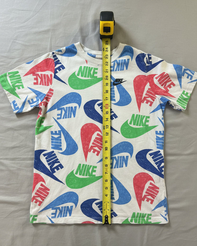A white short-sleeve Nike t-shirt with a colorful all-over logo print, laid flat on a gray textured blanket with a yellow tape measure standing vertically in the center.