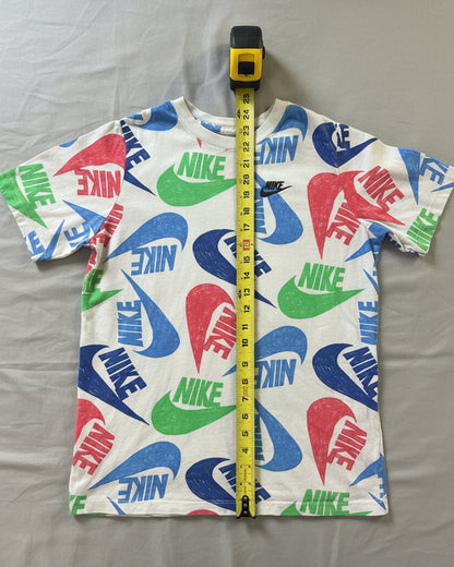 A white short-sleeve Nike t-shirt with a colorful all-over logo print, laid flat on a gray textured blanket with a yellow tape measure standing vertically in the center.