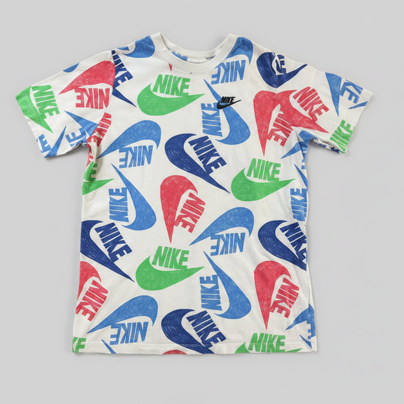 A white short-sleeve Nike t-shirt with an all-over colorful logo print, laid flat on a textured beige blanket. The print features blue, green, and red swooshes and text.