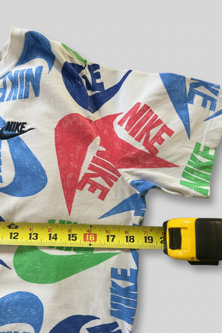 A white Nike t-shirt with a colorful all-over print of blue, red, and green swooshes and text, laid flat on a gray textured blanket with a yellow tape measure showing the size.