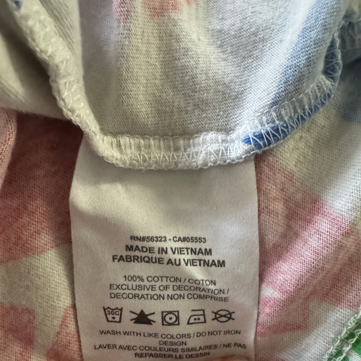 Close-up of a clothing care label reading 'MADE IN VIETNAM' and '100% COTTON', held by fingers against a soft pink, white, and green patterned fabric background.