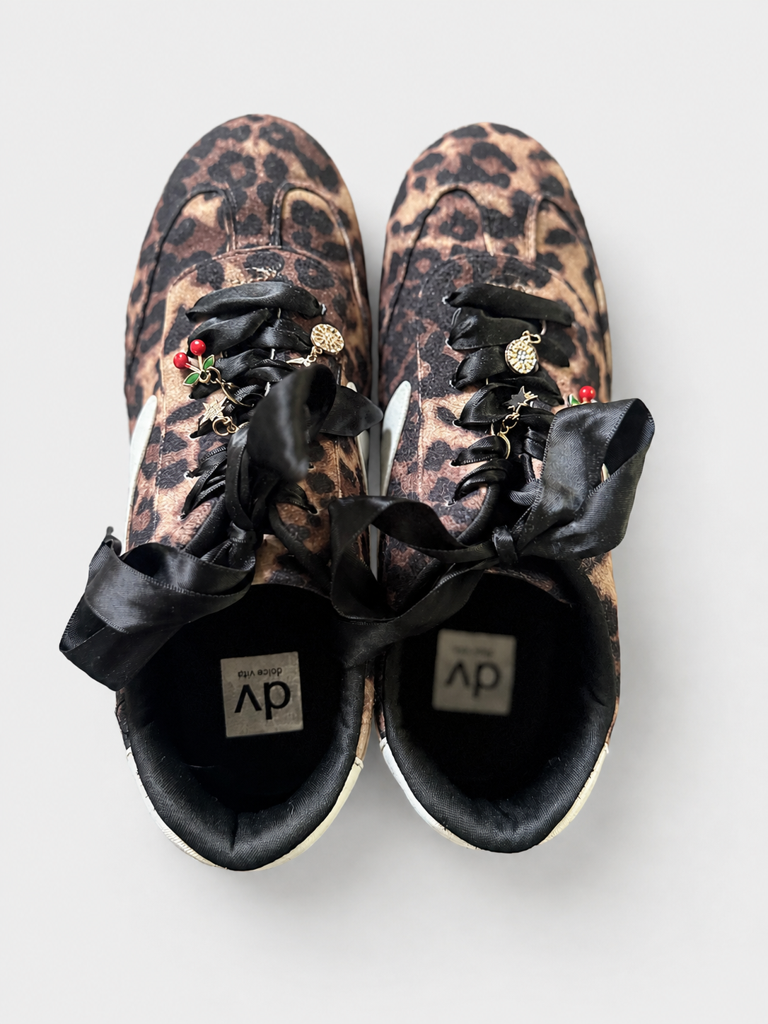 A pair of leopard-print sneakers with black satin ribbon laces adorned with small charms, shown from a top-down view on a light gray background.