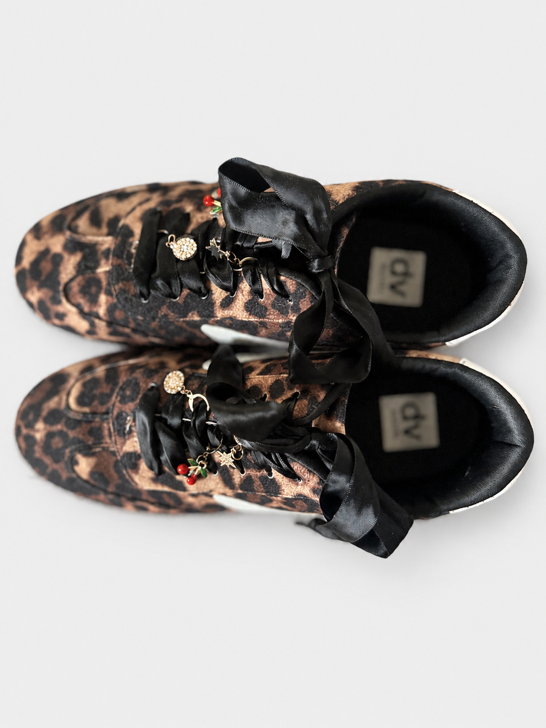 A pair of low-top sneakers with a brown and black leopard print pattern, featuring oversized black satin ribbon laces adorned with small gold charms, photographed from a top-down angle on a light gray background.