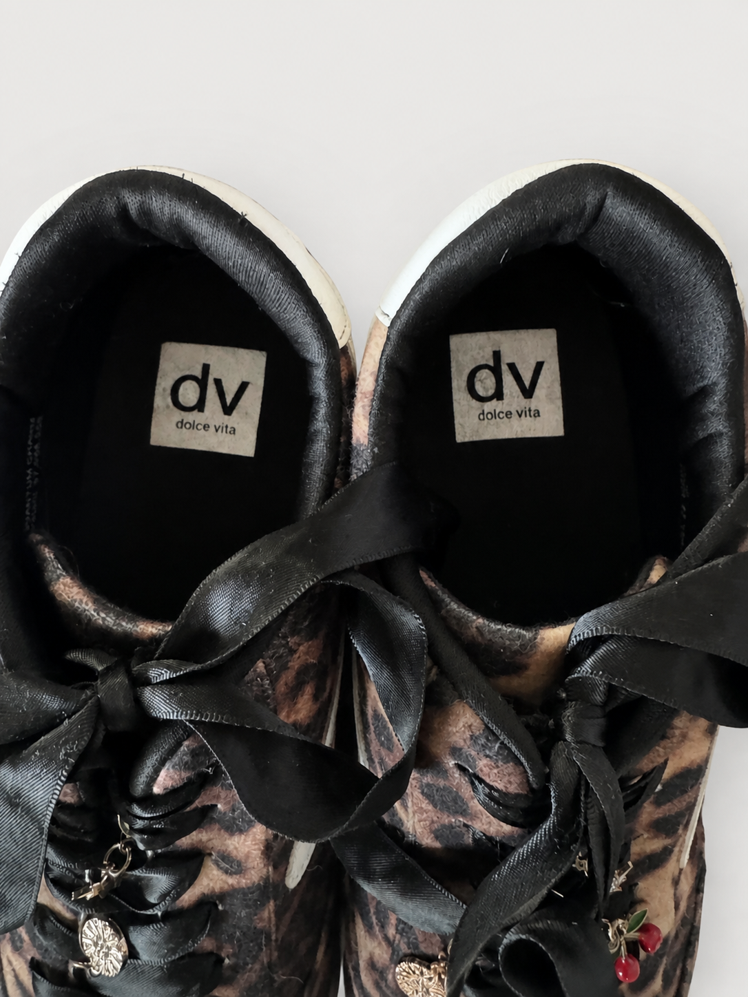 A pair of Dolce Vita low-top sneakers featuring a brown and black animal print pattern, black satin ribbon laces, and small cherry charms, shown from a top-down angle on a light gray background.