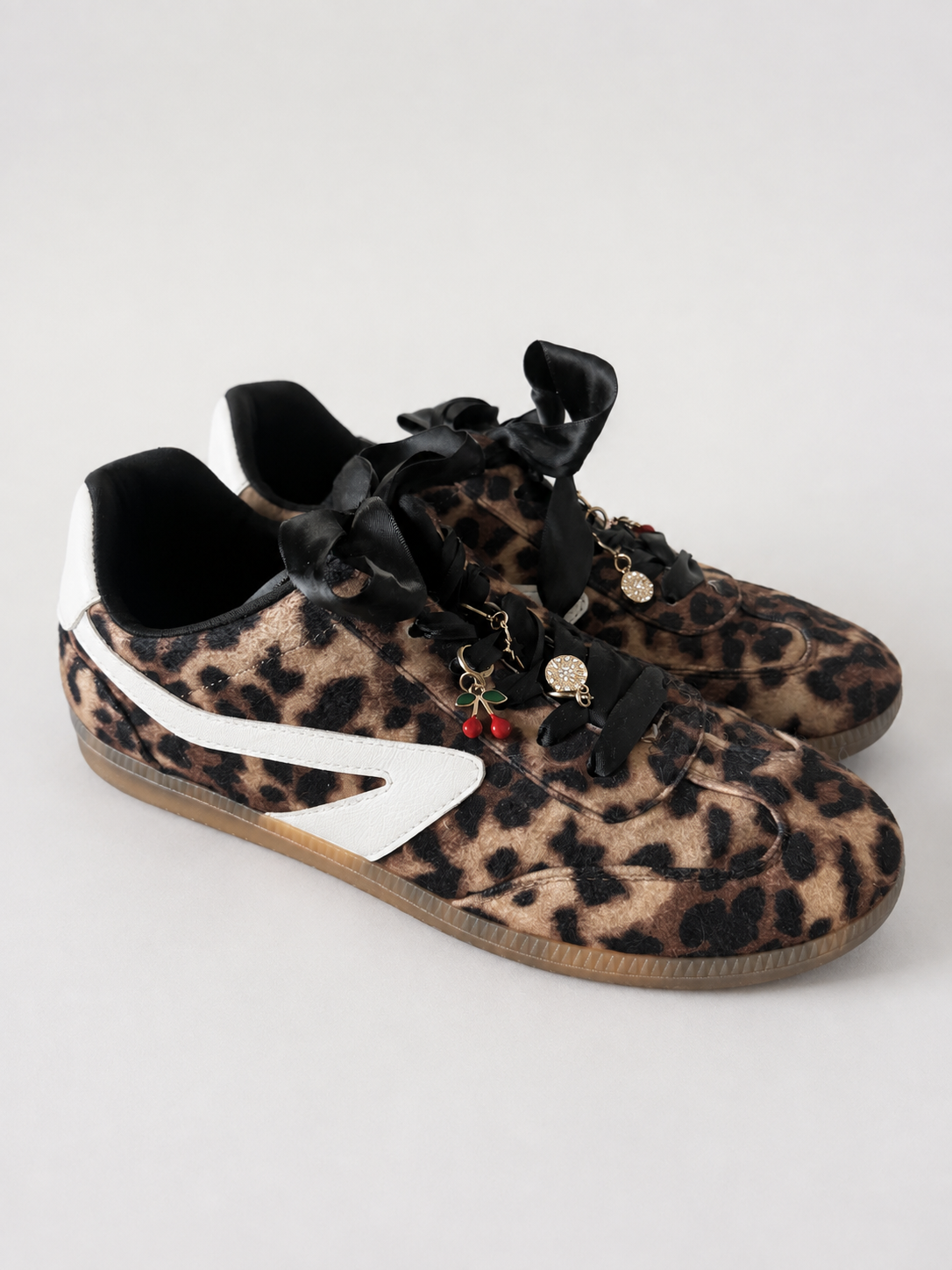 A pair of low-top sneakers with a leopard print pattern, featuring black ribbon laces, small hanging charms, a white side stripe, and brown gum soles, photographed on a light gray background.