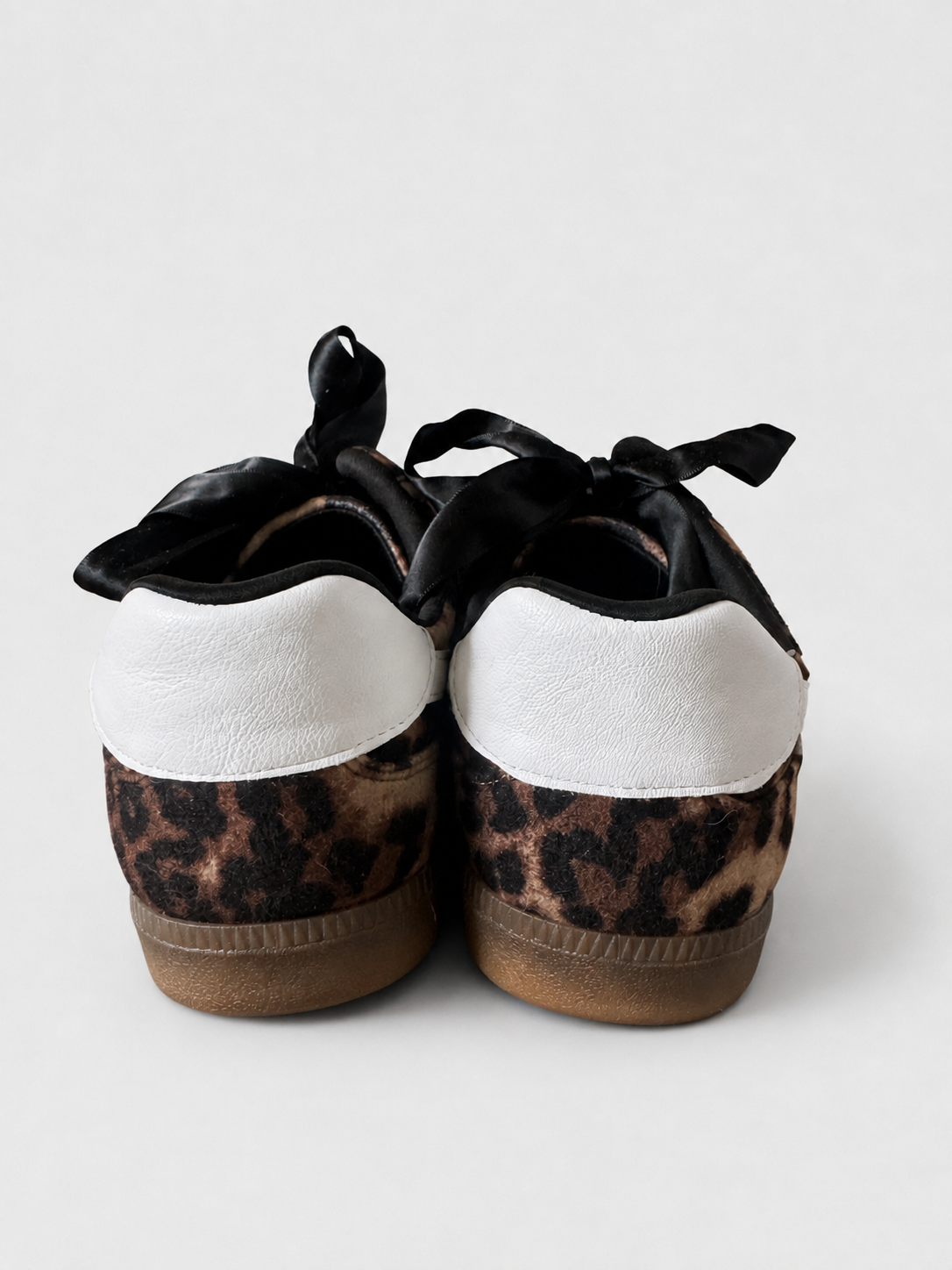 A pair of low-top slip-on shoes shown from the back, featuring a leopard print pattern on the lower half, a white upper section, black satin laces, and brown rubber soles, set against a light gray background.