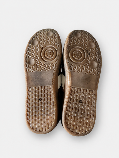 The soles of a pair of brown shoes, showing textured tread patterns and a circular logo design, photographed from a top-down view on a light gray background.
