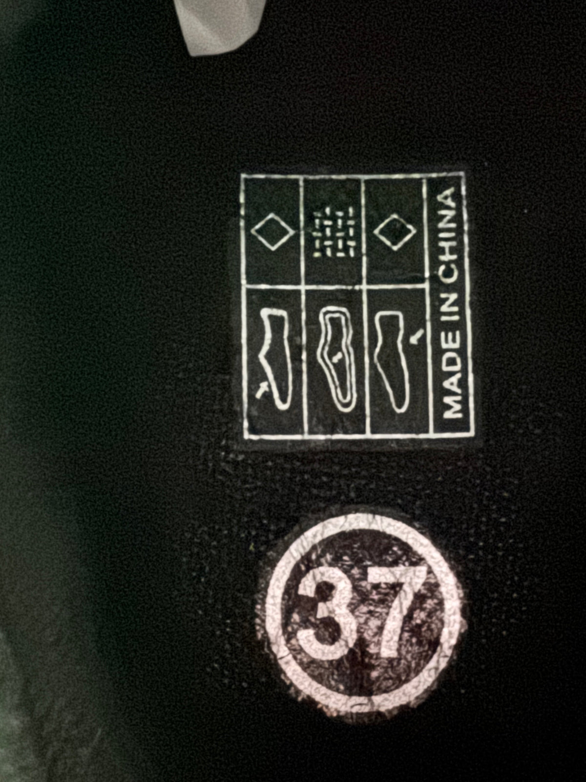 Close-up of a black shoe interior label showing a circular size stamp with the number 37 and a square 'MADE IN CHINA' compliance patch with shoe silhouette icons.