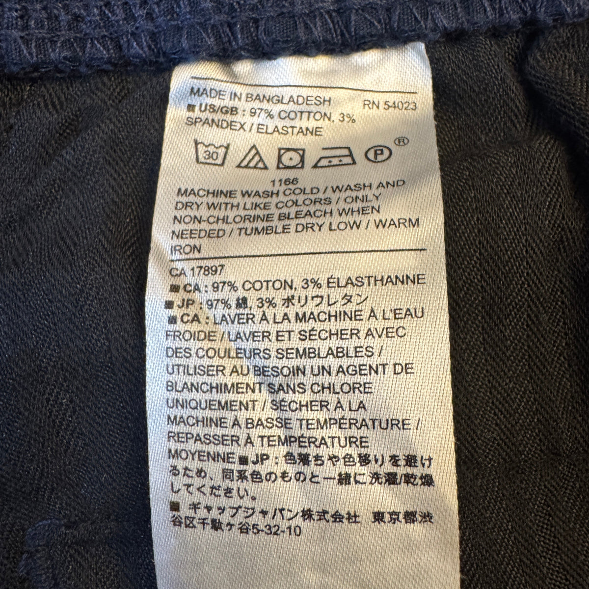 Close-up of a sewn-on fabric care label on dark navy or black knit clothing, showing text including 'MADE IN BANGLADESH', 'COTTON 97%, ELASTHAN 3%', and care symbols.
