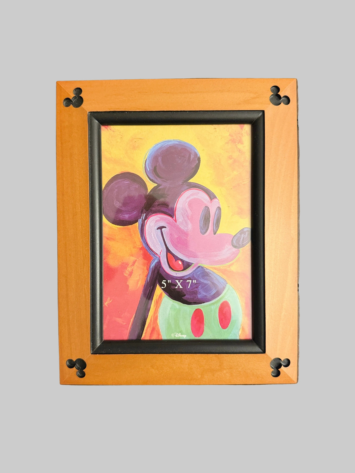 A framed painting of Mickey Mouse on a warm wood table. The artwork features a colorful, textured painting of Mickey with a yellow and orange background, enclosed in a light orange-brown wooden frame with black accents and Mickey-shaped corner details.