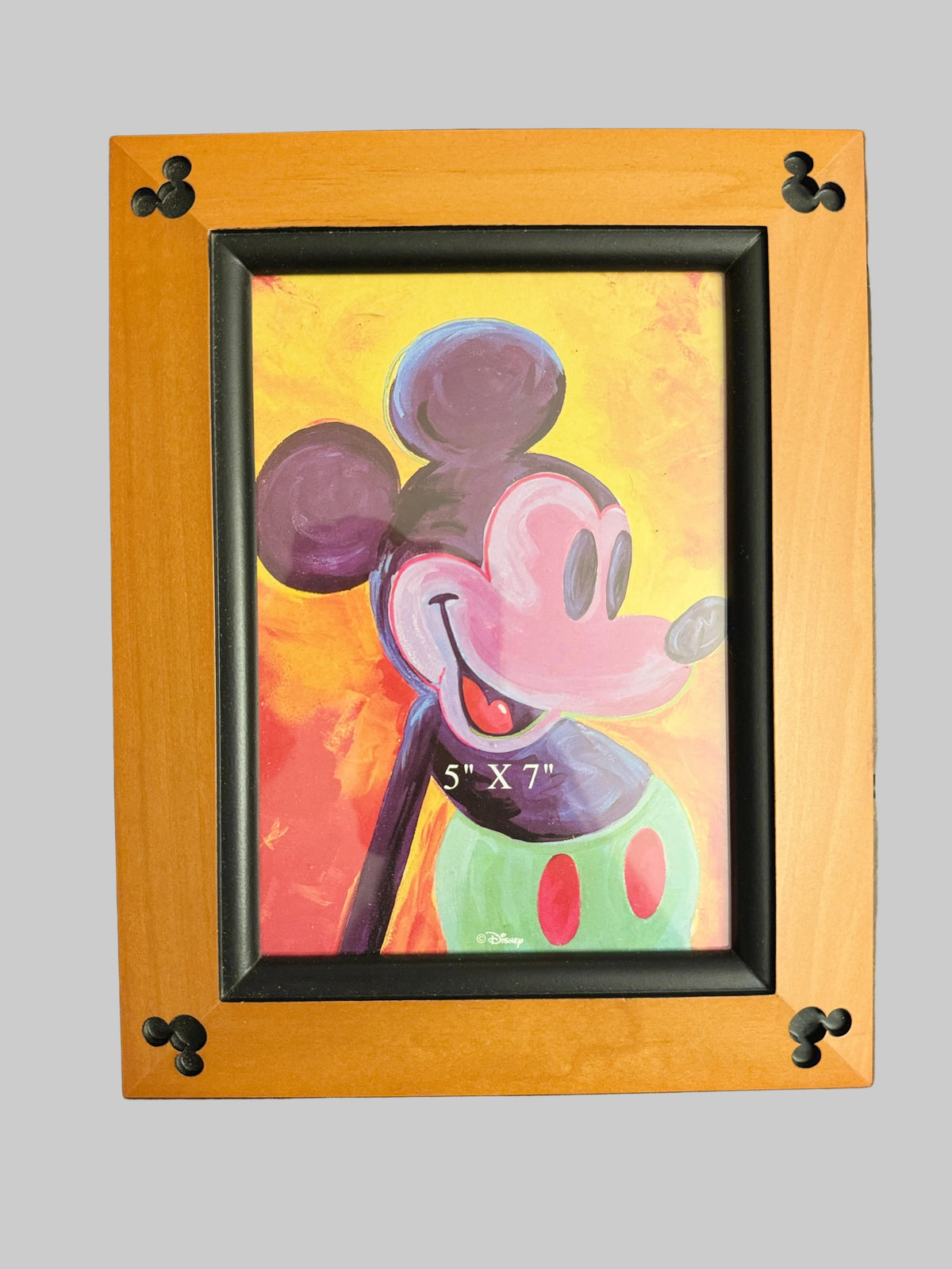 A framed painting of Mickey Mouse on a wooden table. The artwork features a colorful, textured painting of Mickey with a purple head, pink face, and green shorts against a yellow and orange background.