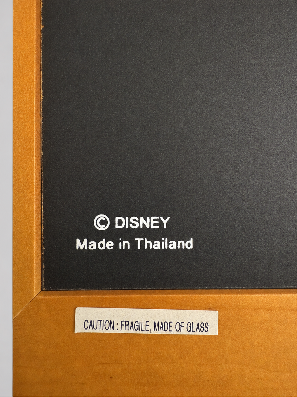 Close-up of a Disney framed art piece showing a black center panel with white 'Disney Made in Thailand' text, a white caution label reading 'CAUTION: FRAGILE, MADE OF GLASS,' and a warm orange-brown wooden frame.