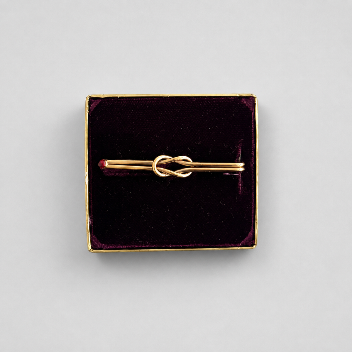A gold-toned pin with a central knot design, presented in a square dark velvet box with a gold border on a light gray background.
