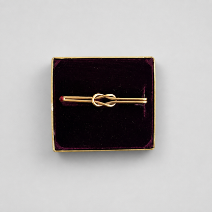 A gold-toned pin with a central knot design, presented in a square dark velvet box with a gold border on a light gray background.