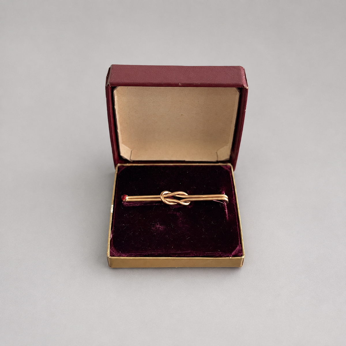 A gold tie-clasp pin with a small red gemstone on each end, presented in an open burgundy velvet jewelry box on a neutral gray background.