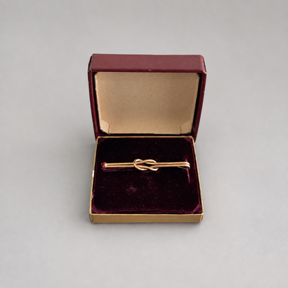 A gold tie-clasp pin with a small red gemstone on each end, presented in an open burgundy velvet jewelry box on a neutral gray background.