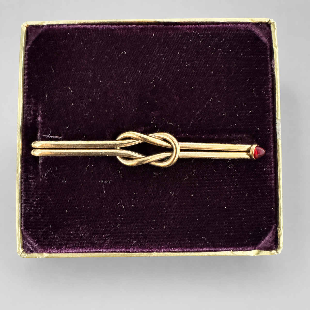 A gold tie bar with a knotted center design and a small red gemstone accent on one end, presented in a square dark purple velvet jewelry box.