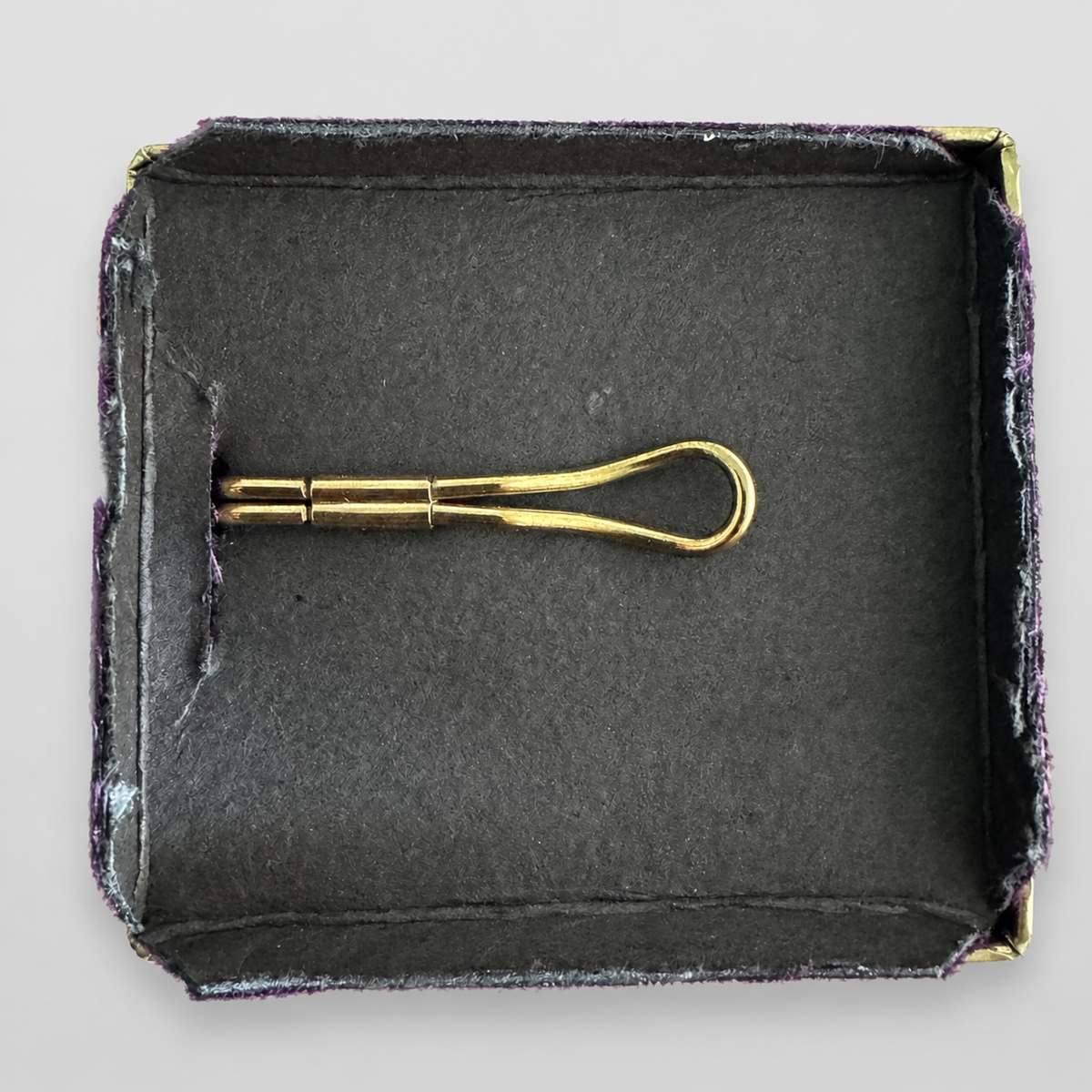A gold metal tie clip displayed inside an open dark gray velvet presentation box on a light gray background.