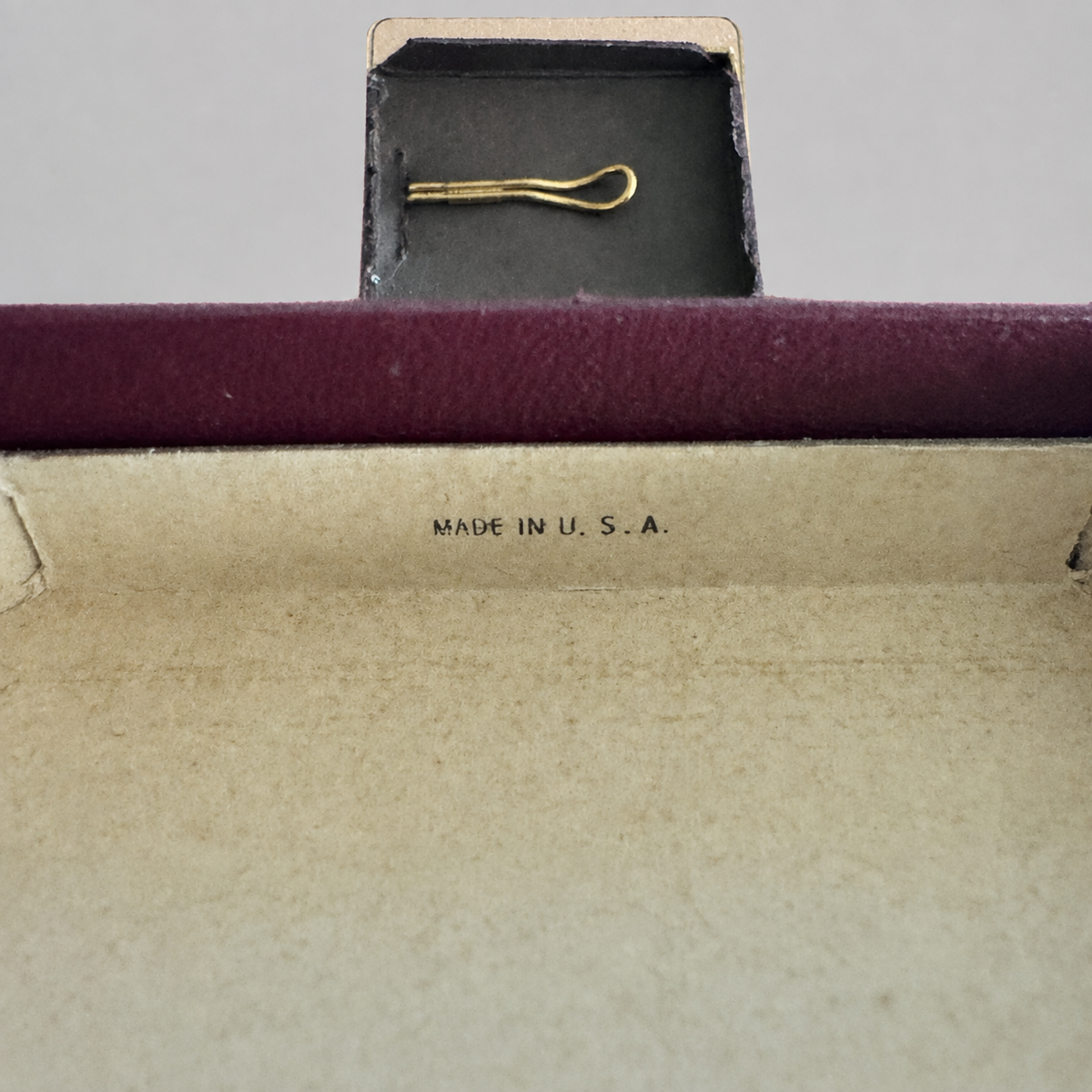 A close-up of a vintage gold hair tie or barrette displayed inside an open dark plum velvet jewelry box with a beige interior. The text 'MADE IN U.