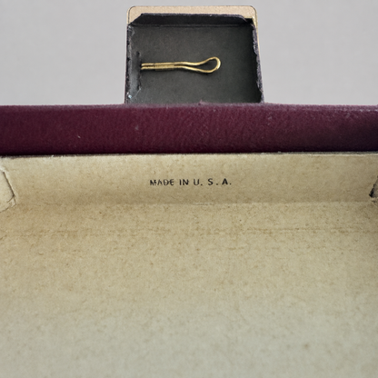 A close-up of a vintage gold hair tie or barrette displayed inside an open dark plum velvet jewelry box with a beige interior. The text 'MADE IN U.