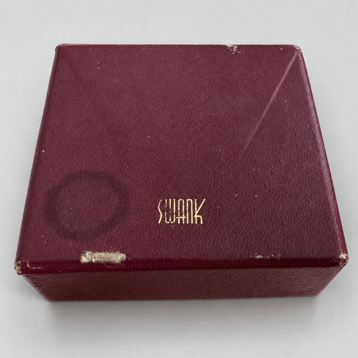 A vintage Swank brand burgundy textured box with gold lettering, photographed on a neutral light gray background.