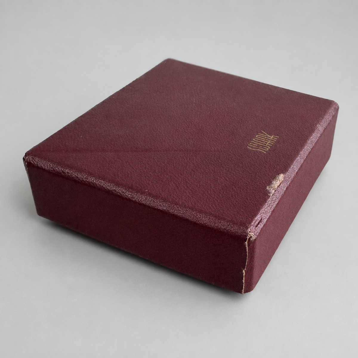 A square vintage watch box made of textured burgundy leather with gold foil lettering on the lid, photographed on a neutral gray background.