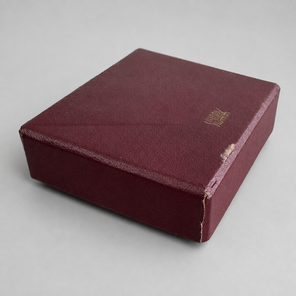 A square vintage watch box made of textured burgundy leather with gold foil lettering on the lid, photographed on a neutral gray background.