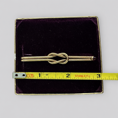 A gold-toned metal pin with a knotted bar design, displayed inside an open dark purple velvet jewelry box with a yellow Stanley tape measure placed vertically beside it for scale, on a light gray background.