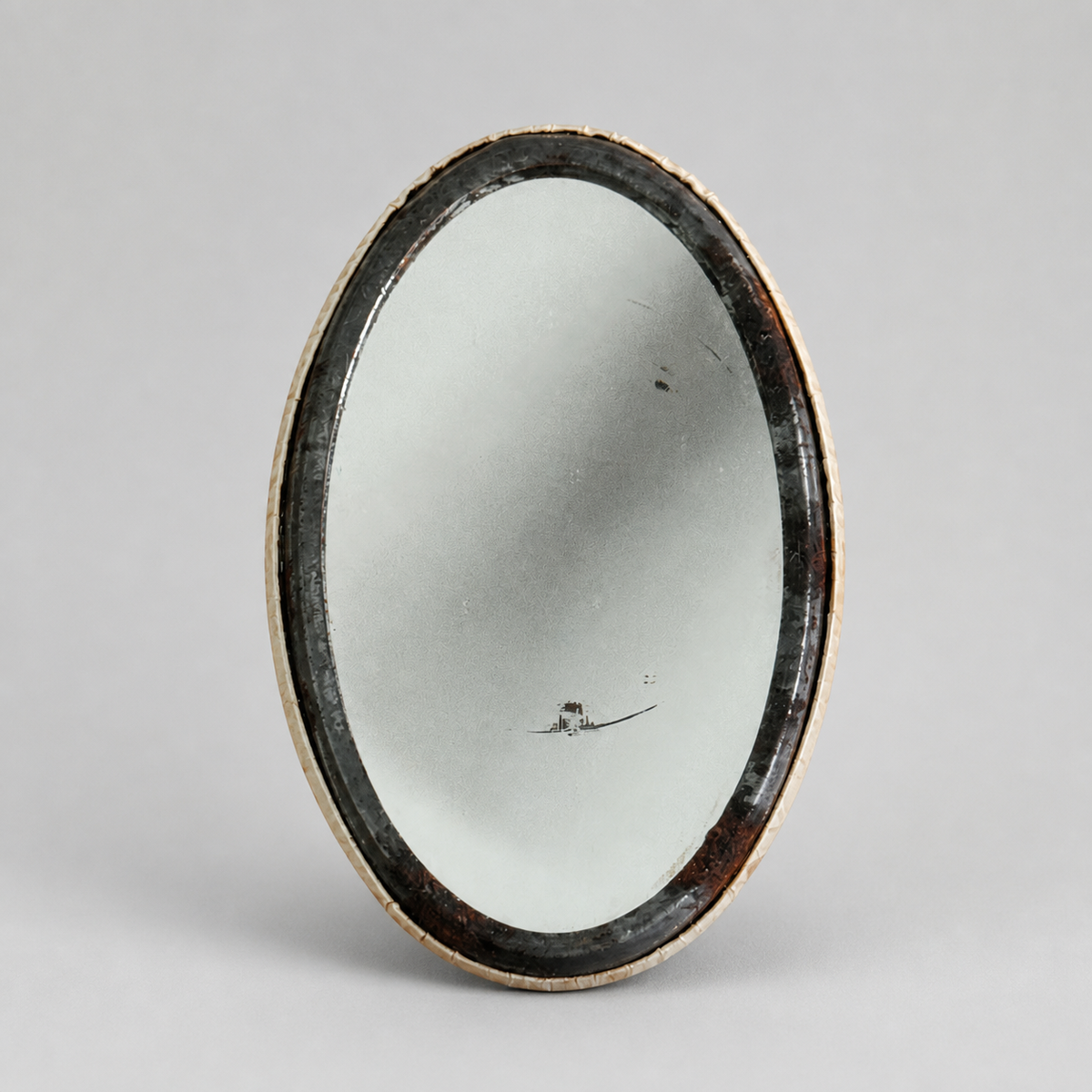 An oval vintage mirror with a dark distressed frame, standing upright against a neutral light gray background.