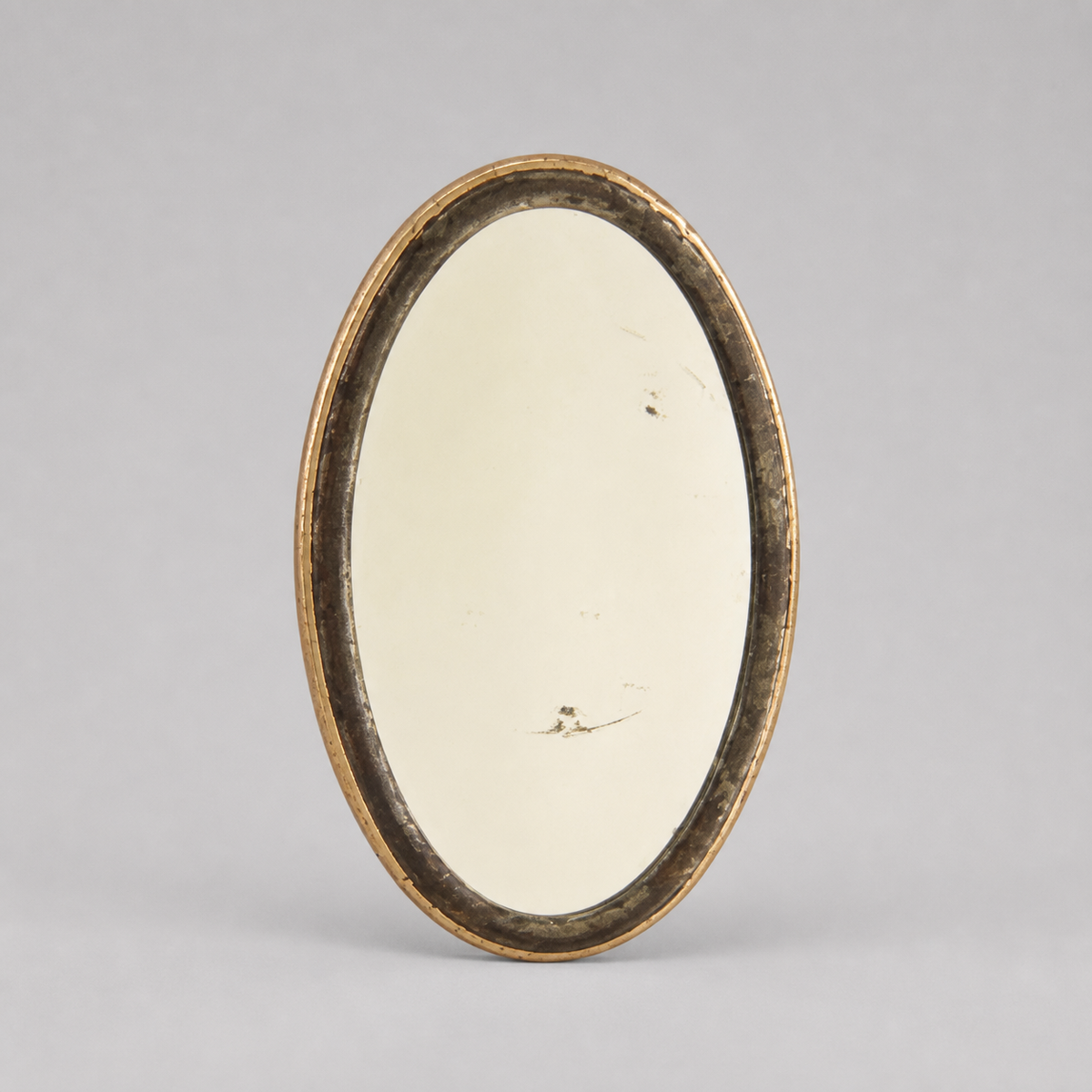 An oval vintage mirror with a distressed gold-toned frame, standing upright against a neutral gray background.