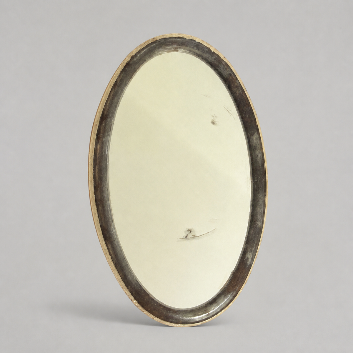 An oval vintage mirror with a dark, distressed frame and a gold-toned edge, standing upright against a neutral gray background.
