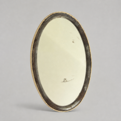 An oval vintage mirror with a dark, distressed frame and a gold-toned edge, standing upright against a neutral gray background.