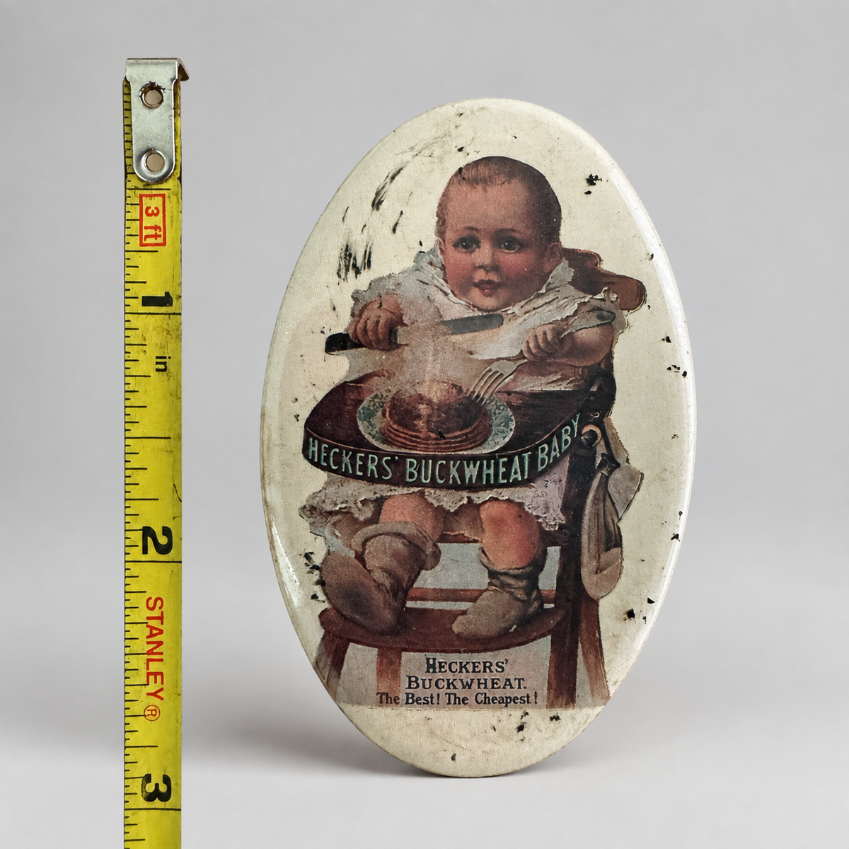 A vintage oval tin sign featuring a baby in a high chair with the text 'Heckers' Buckwheat Baby,' shown next to a yellow Stanley tape measure on a light gray background.