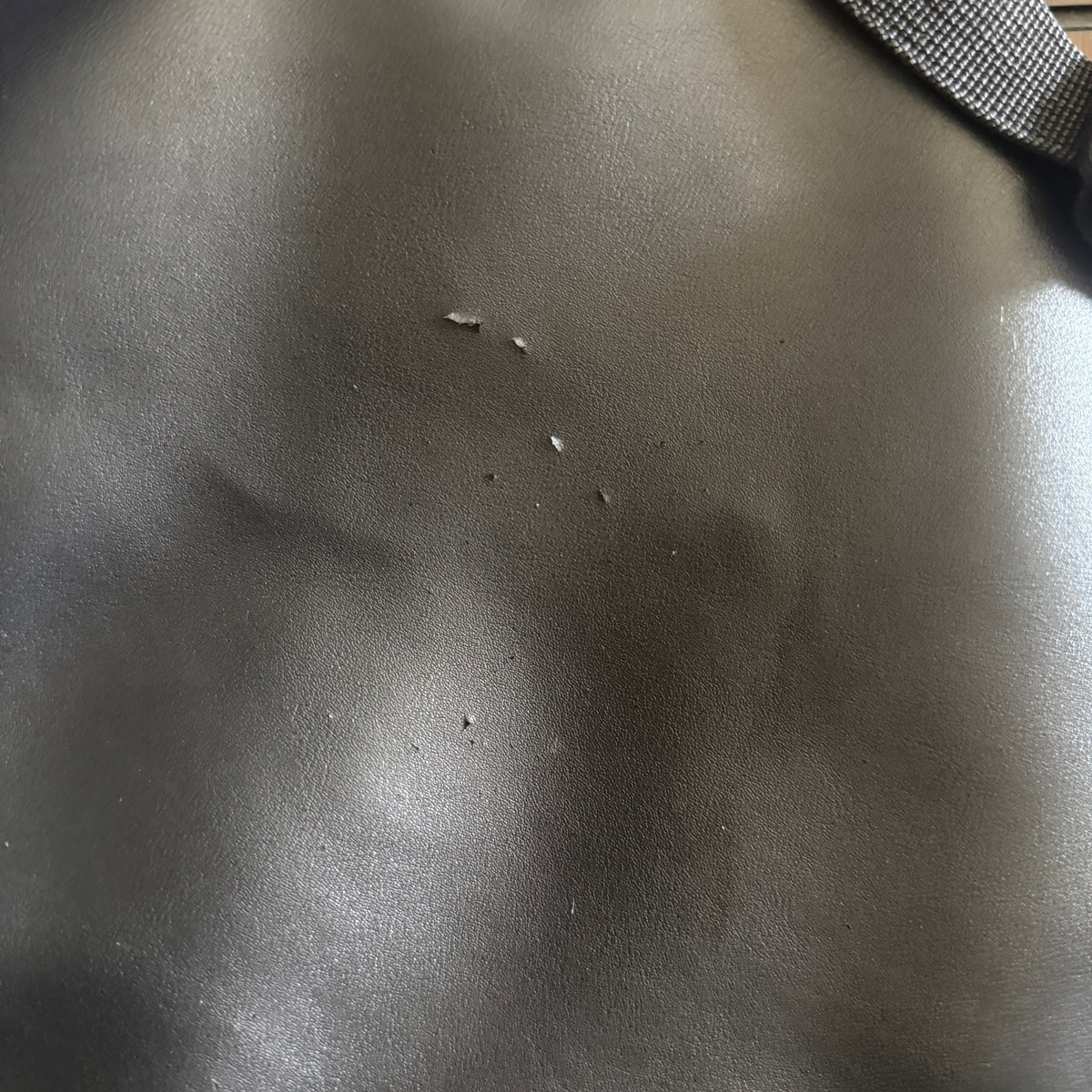 Close-up of a dark gray textured leather or faux-leather surface with a few small scuffs and a gray woven strap visible in the top right corner.