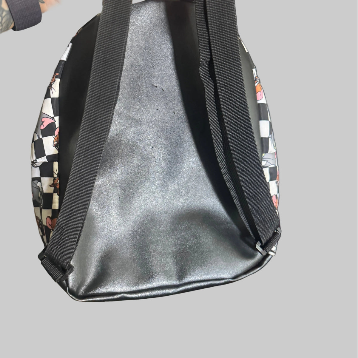 A black faux-leather tote bag with black shoulder straps, held by a tattooed hand. The sides of the bag feature a black and white checkerboard pattern with small red and white graphic details.