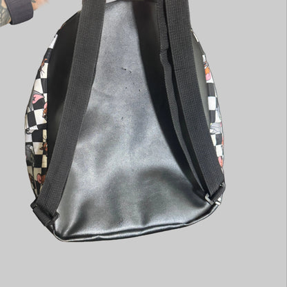 A black faux-leather tote bag with black shoulder straps, held by a tattooed hand. The sides of the bag feature a black and white checkerboard pattern with small red and white graphic details.