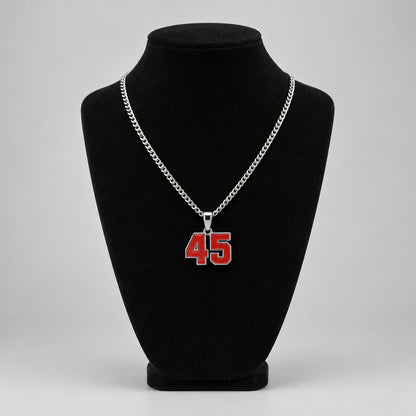 A silver chain necklace with a red and white '45' number pendant displayed on a black velvet mannequin bust. A tattooed hand is visible holding the necklace above the bust.