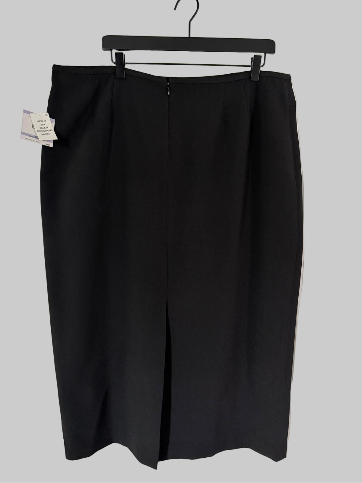 A black knee-length skirt hanging on a black hanger against a white paneled door, with a small product tag still attached on the left side.