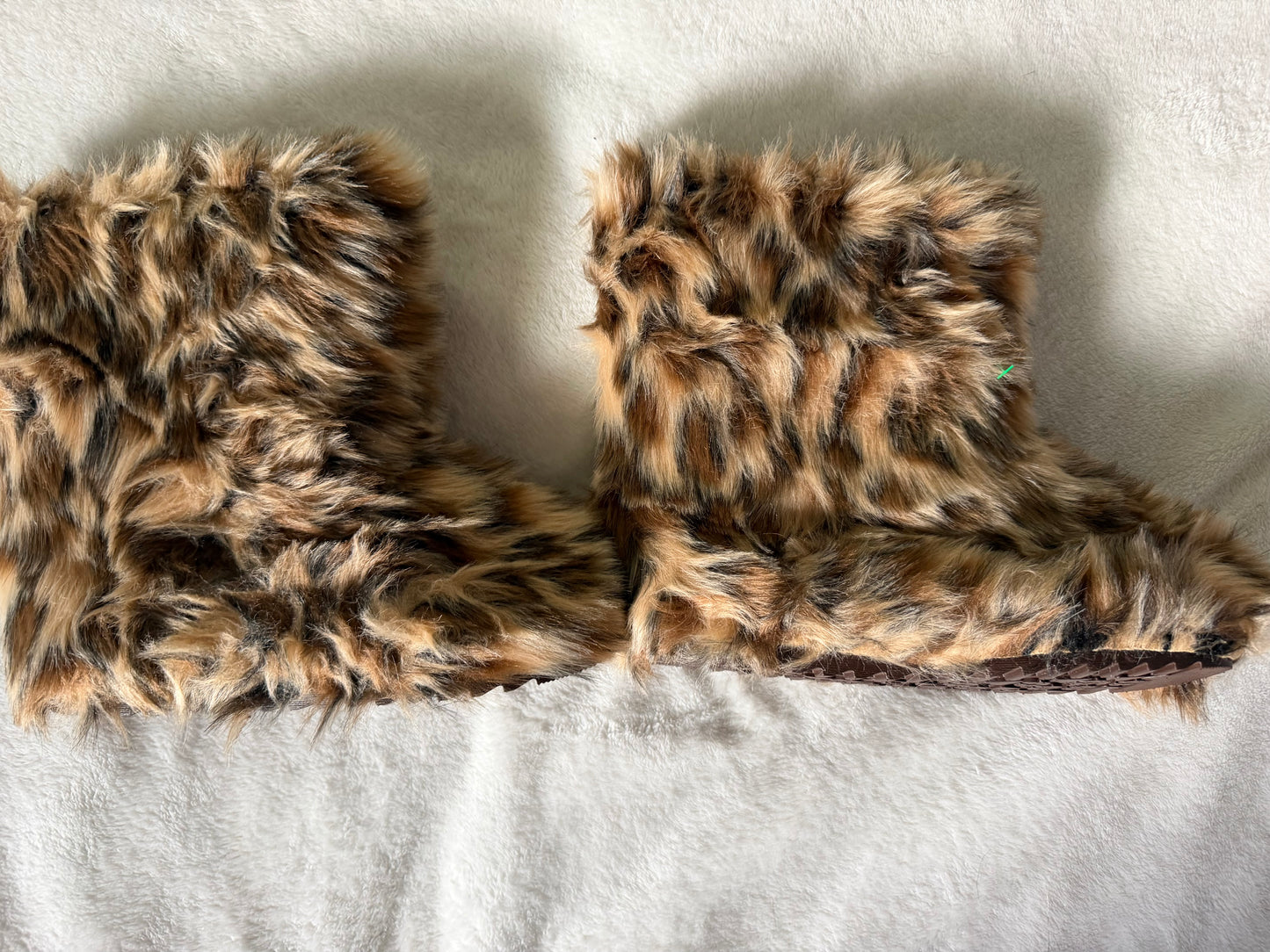 A pair of plush faux-fur slipper booties in a brown, black, and beige leopard print, laid flat on a white textured fabric background.