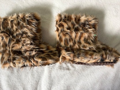A pair of plush faux-fur slipper booties in a brown, black, and beige leopard print, laid flat on a white textured fabric background.