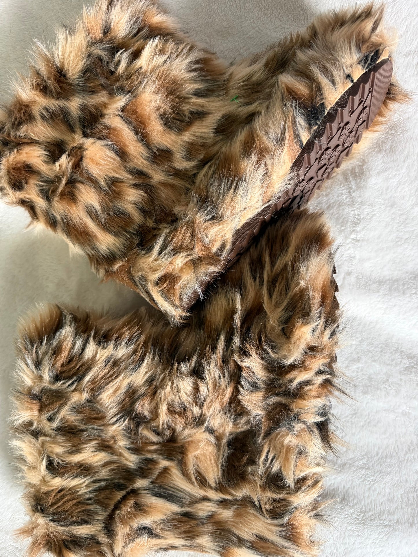 A pair of plush faux-fur slippers in a brown, black, and tan leopard print pattern, shown on a white textured fabric background with one slipper turned to show the sole.