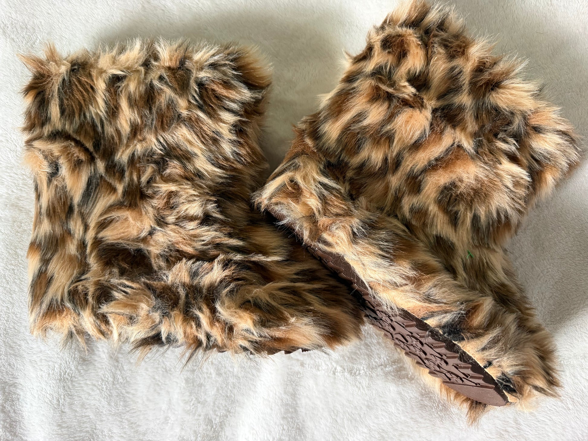 A pair of brown and black faux fur wrist warmers with a brown woven strap, laid flat on a white textured fabric background.