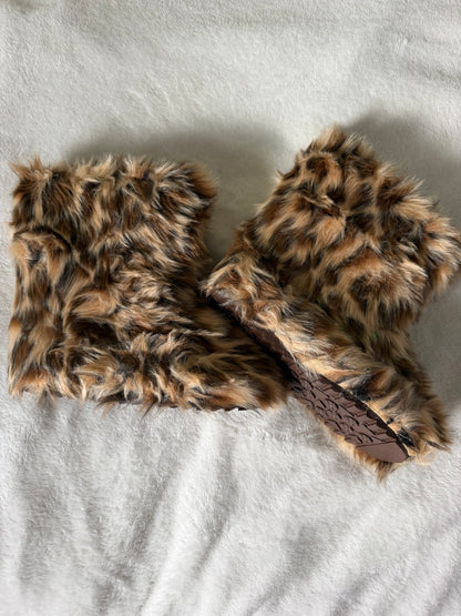 A pair of brown and black faux fur slippers with a brown textured sole, laid flat on a white textured fabric background.