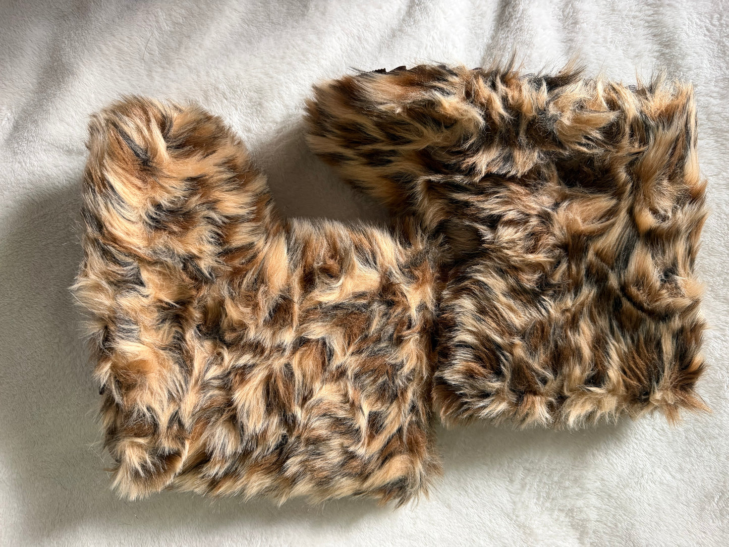 A pair of faux fur boot covers in a brown, black, and beige leopard print pattern, laid flat on a white textured fabric background.