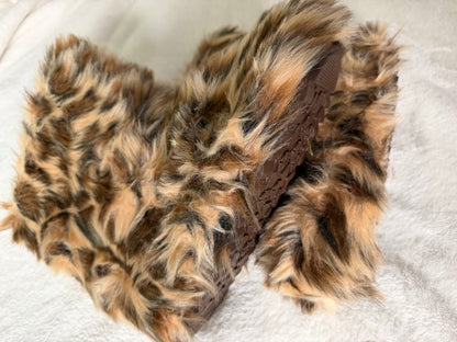 A pair of fuzzy faux-fur slippers in a brown, beige, and black leopard print pattern, with a brown textured sole, resting on a white textured blanket.