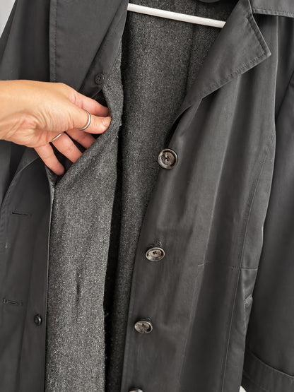 Close-up of a dark charcoal gray coat with a hand holding open the front to reveal a textured gray wool-like lining, shown on a hanger.